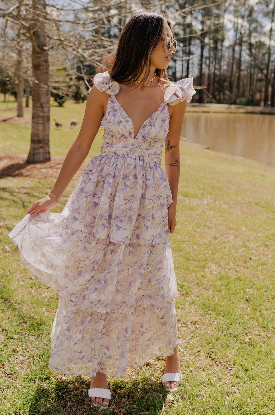 front view of model wearing The Harlow Lavender Floral Ruffle Midi Dress features sheer fabric with purple, green, orange floral print, shimmer thread details, beige lining, a tiered body with a maxi-length hem, ruffled straps, a plunge neckline, a lace-up back, and a back zipper and hook closure.