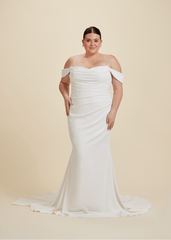 A woman smiles with her hands on her hips, wearing the Luxe Privée Louella off-the-shoulder crepe fit and flare wedding dress by PLR, featuring a ruched bodice, posed against a plain beige background.