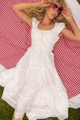 A woman in the Ivy City Co Palermo Dress in White and red sunglasses lies relaxed with one arm above her head on a red and white checkered picnic blanket spread on the grass.