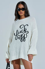With long dark hair and sunglasses, a person wears the EFF OFF KNIT SWEATER DRESS, an oversized, relaxed-fit white sweater with a bold black cursive phrase. They hold a strap against a neutral background, exuding an effortlessly chic vibe.