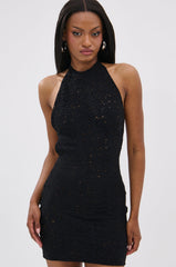A woman faces forward with a neutral expression against a plain white background, wearing the ON THE MOVE BODYCON MINI DRESS IN BLACK—a sleeveless halter neck mini dress featuring openwork crochet details.