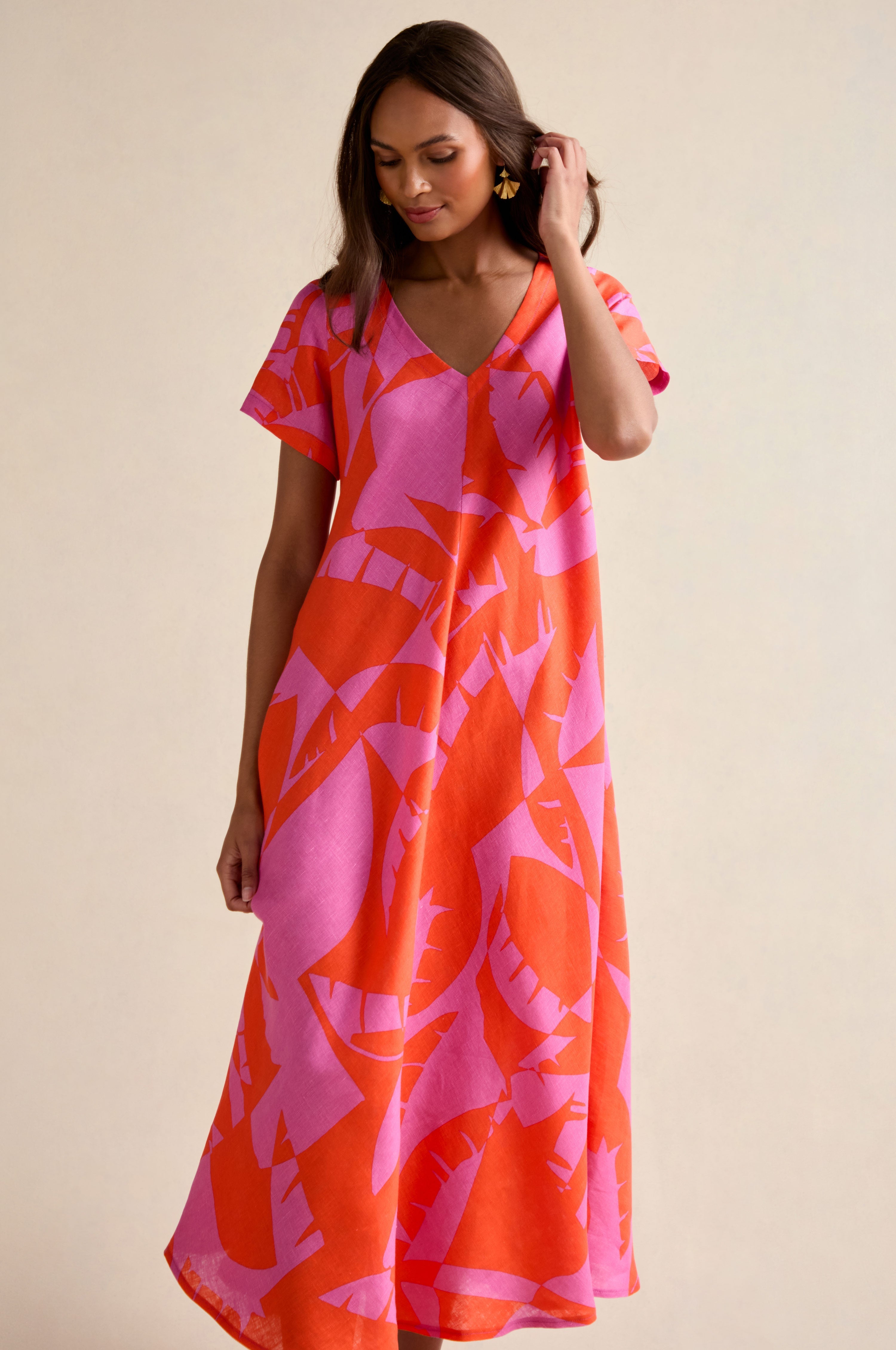 A woman wears the Palm Mirage Linen Flared Dress, featuring a flowing, short-sleeved design with a red and pink abstract leaf pattern, standing against a neutral background.