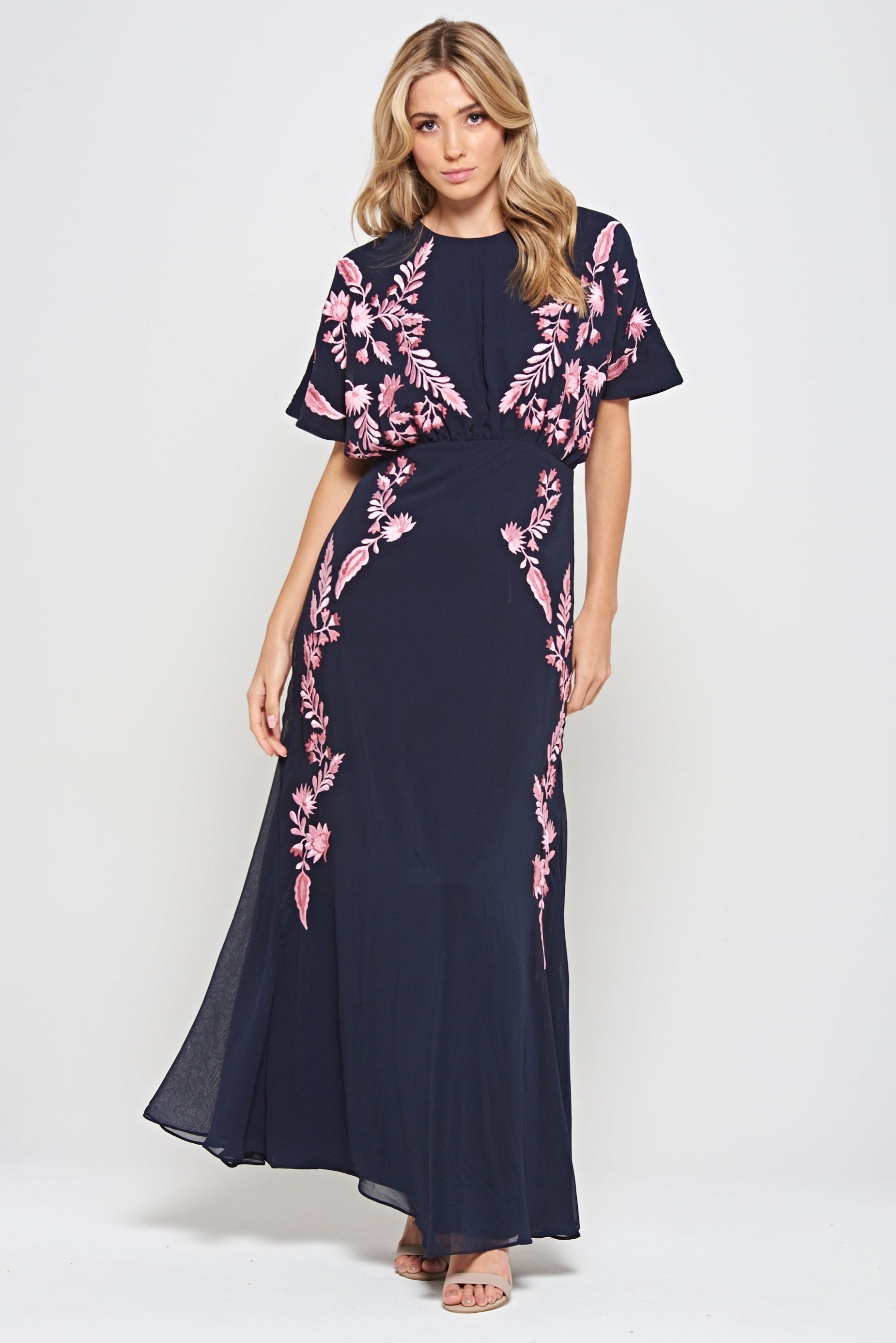 Navy floral embroidered maxi dress with pink detailing, worn by a model. Features flutter sleeves, a fitted waist and a flowing full-length skirt—perfect for weddings, evening occasions and special events.