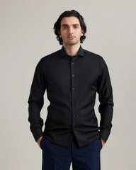 Man wearing a black merino dress shirt from Wolk and navy pants against a plain background