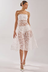 A woman showcases the MAYA ABSTRACT MIDI DRESS IN WHITE, a strapless, sheer midi with abstract swirls and a flared skirt. She elevates her cocktail look with white heels, gold earrings, and sleek, pulled-back hair.