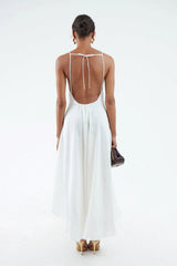 Back view of a white, maxi silk-hemp dress featuring thin shoulder straps that extend down the length of a low open back. Straps are connected across the upper back with a horizontal tie. Low back edge has elastic waist gather, giving dress an airy and flowing silhouette. Model holding maroon clutch.