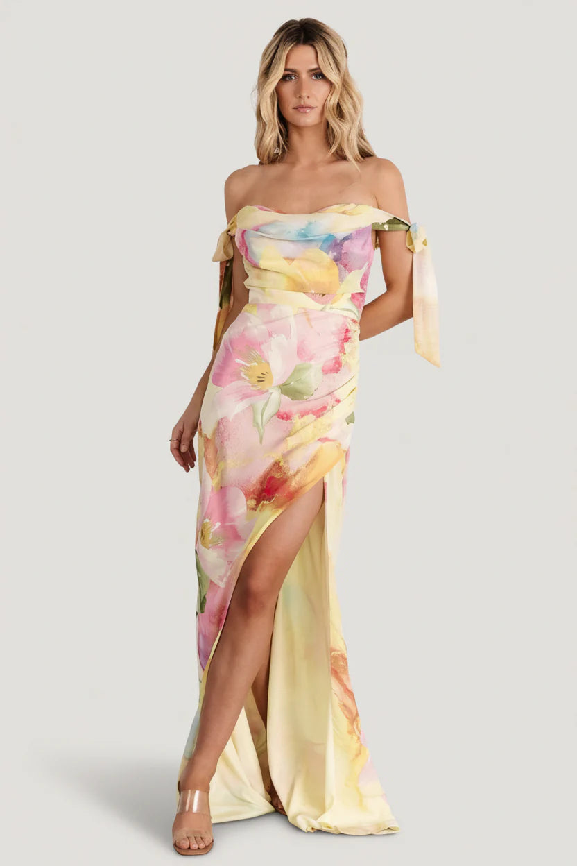 Front view of yellow multi-floral off-shoulder chiffon maxi dress showing cowl neckline, adjustable bow straps, and tulip skirt in motion.