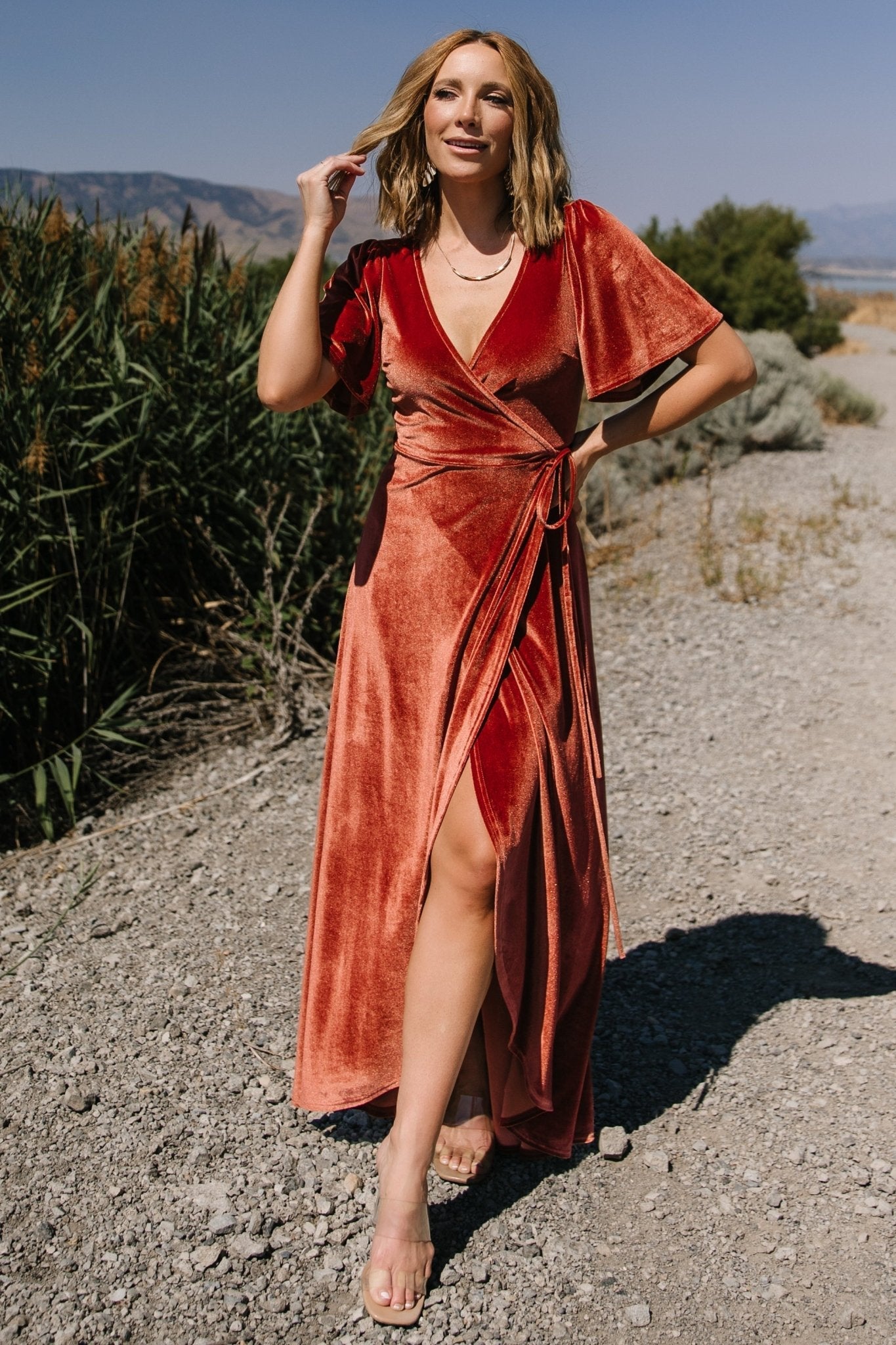 Front view of Cinnamon Katelyn velvet maxi wrap dress on model showing V-neckline, tulip skirt and medium flutter sleeves, highlighting fabric sheen and drape.
