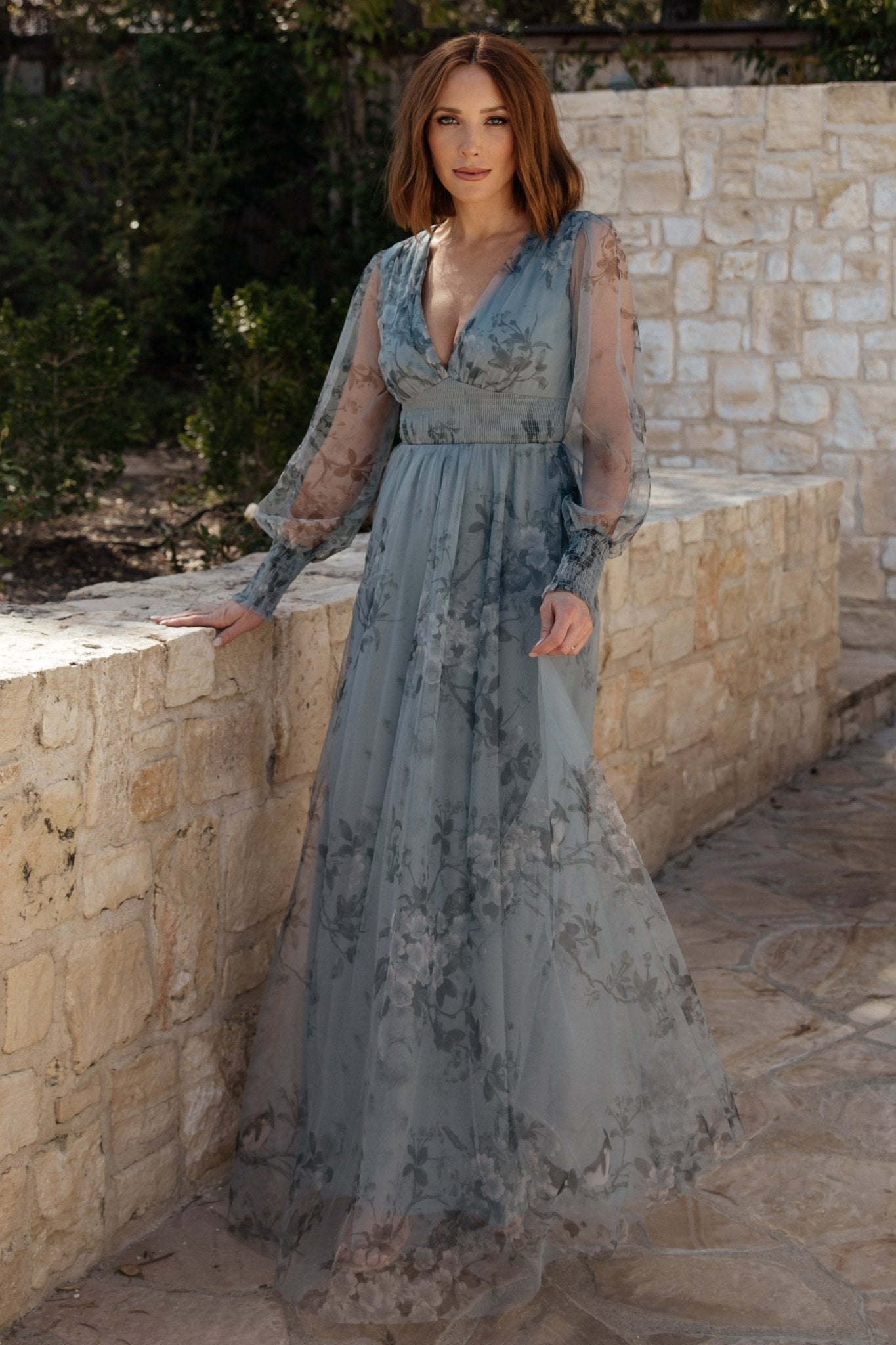 Front view of dusty blue floral tulle maxi dress with deep V-neck, smocked waist, long sheer sleeves, and flowing skirt, model standing.