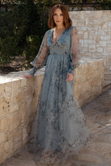 Front view of dusty blue floral tulle maxi dress with deep V-neck, smocked waist, long sheer sleeves, and flowing skirt, model standing.