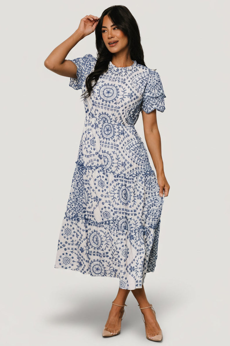 Front view of white midi dress with blue embroidered eyelet, tiered midi skirt, high ruffle neckline, scalloped short sleeves.