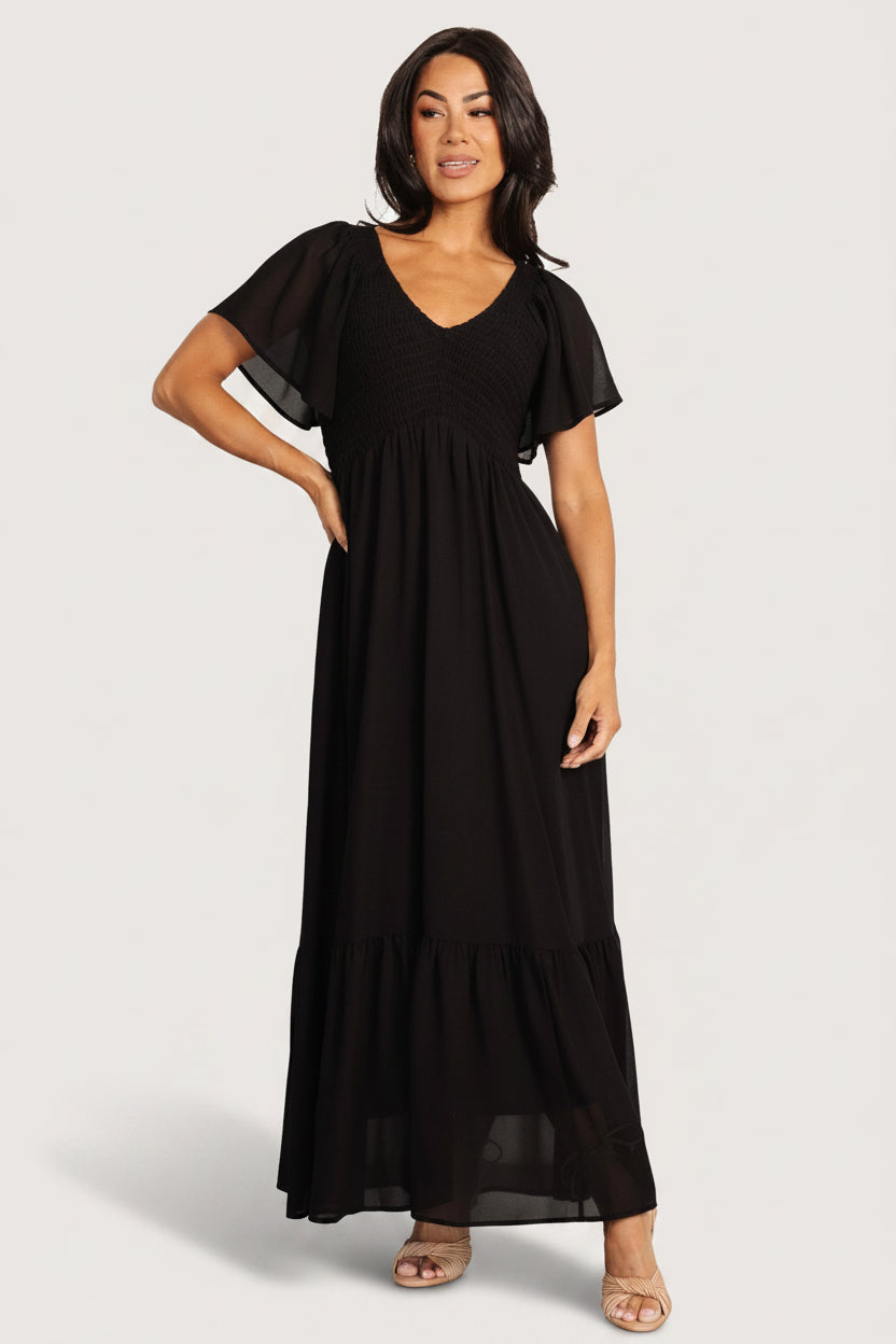 Front view of Monica Smocked Maxi Dress in black chiffon showing V-neckline, center-seamed smocked bodice, flutter sleeves, and flowing maxi skirt.