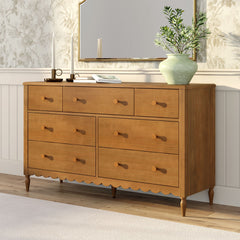 Light oak solid wood dresser with multiple drawers and scalloped apron, styled with green ceramic vase, books and candles under a gold-framed mirror in a bright farmhouse bedroom — ideal bedroom storage and home decor