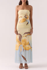 A woman stands with arms by her sides, wearing the Sovere Dahlia Maxi Dress—a strapless stretch mesh maxi in large yellow and brown florals over a light pastel background—paired with open-toe sandals.