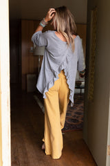 A woman with long hair, barefoot and facing away, walks in a wooden room wearing The Nest Must Have High Low Tee in Heather Gray with wide-legged yellow pants.