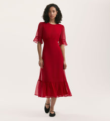 A woman with curly hair wears the Jennifer Red Chiffon Midi Dress, featuring a round neck, sheer short sleeves, and ruffled hem. She stands against a plain white background and pairs the fit and flare dress with black shoes.
