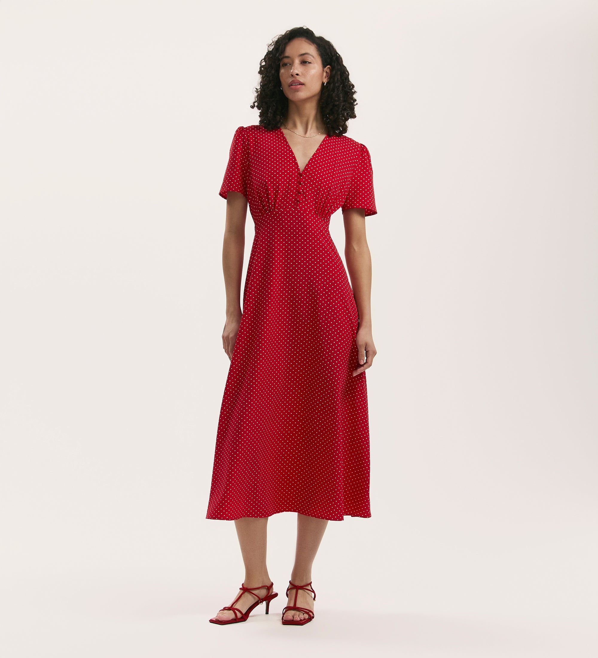 A woman poses against a plain background in the Nieve Red Polka Dot Crepe Midi Tea Dress, featuring short sleeves and a V-neck. The crease-free fabric complements her curly hair, relaxed posture, and red strappy sandals.