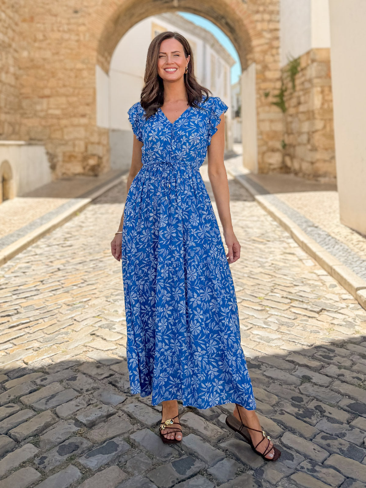 Model wearing a blue floral maxi dress with ruffle cap sleeves and cinched waist, styled with brown sandals on a sunny…