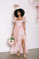 A model wearing a long, pink floral dress with ruffled sleeves and a high-low hemline, holding a bouquet of pink flowers.