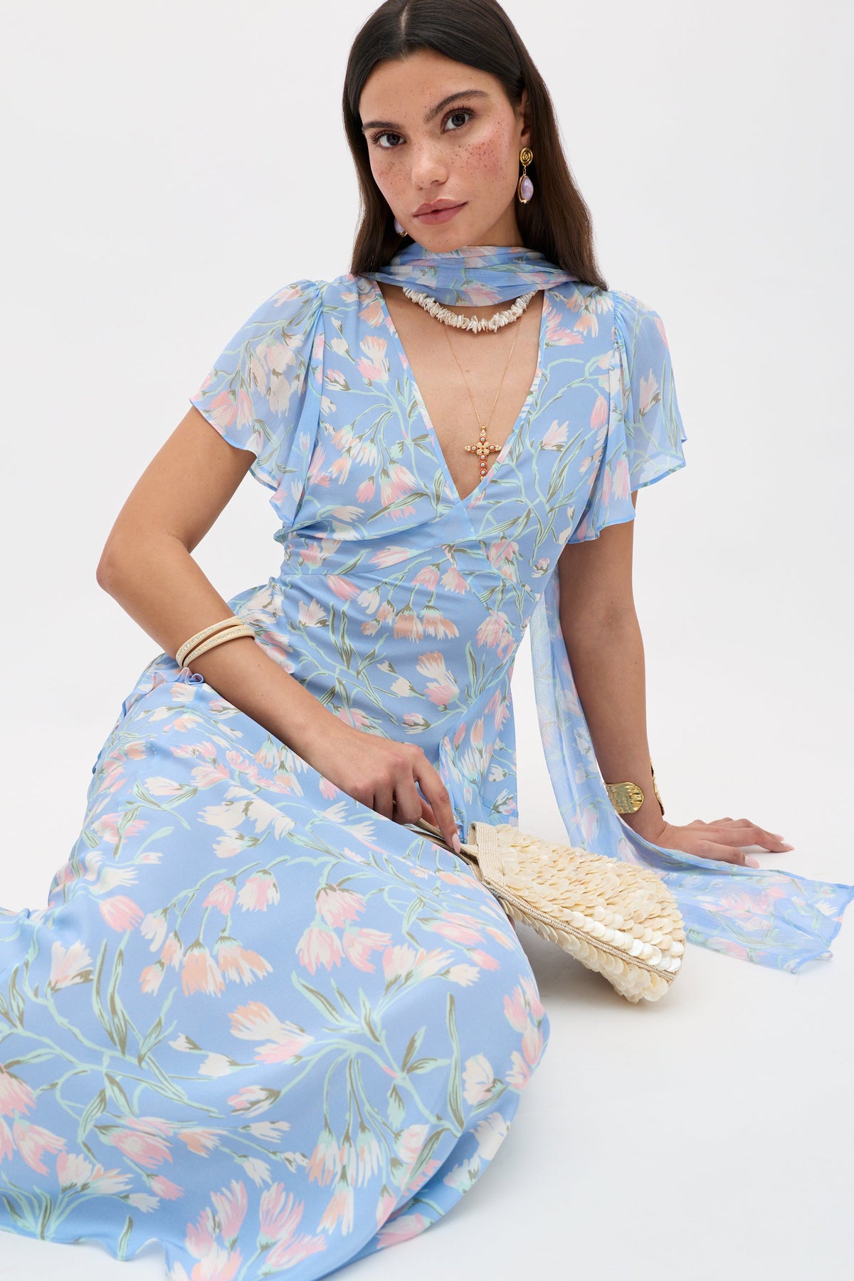 Wearing the Evie - Watercolour Floral Blue dress with a matching scarf, a woman sits on the floor holding a woven fan. She accessorizes with gold jewelry and gazes neutrally at the camera against a plain white background.