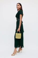 A woman wears the Corrine - Diana Embellishment Forest Green ankle-length dress, paired with a yellow woven handbag, gold open-toe heels, hoop earrings, and a bracelet, standing against a plain light background.