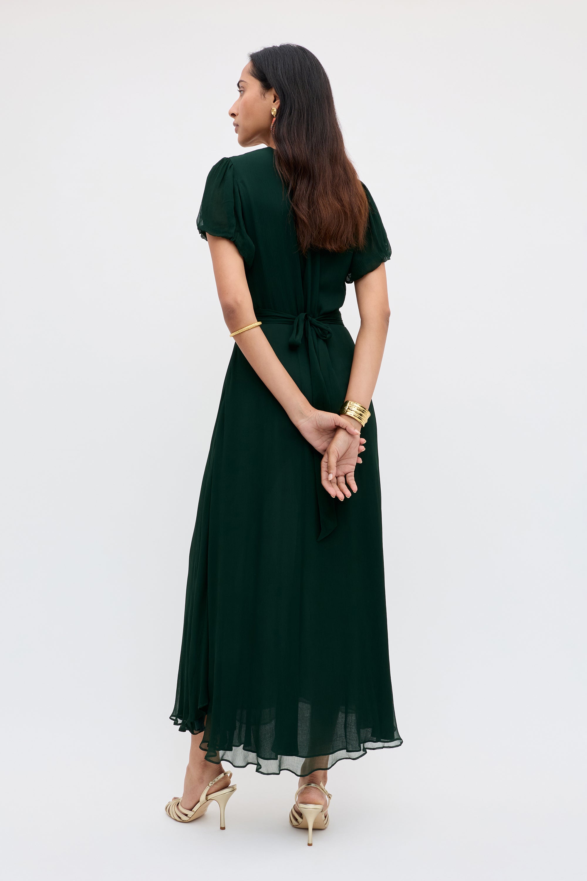 A woman with long brown hair, gold bracelets, and earrings faces away, wearing the Corrine - Diana Embellishment Forest Green dress—an ankle-length, short-sleeve piece with a waist tie—paired with light heeled sandals.