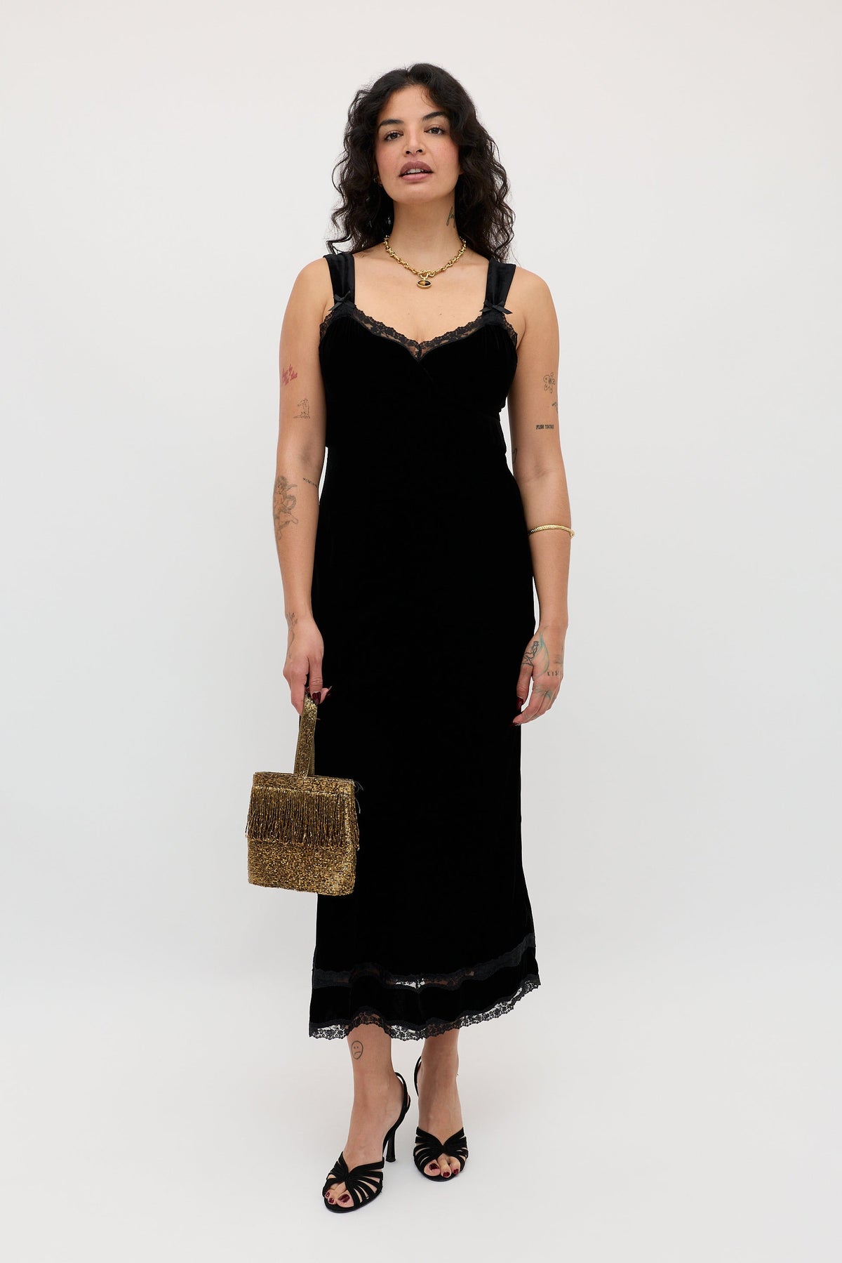 A woman wears the Lucie - Black sleeveless dress with lace trim, accessorized with a woven handbag, black sandals, and gold jewelry. She stands against a plain background, her arm tattoos visible.