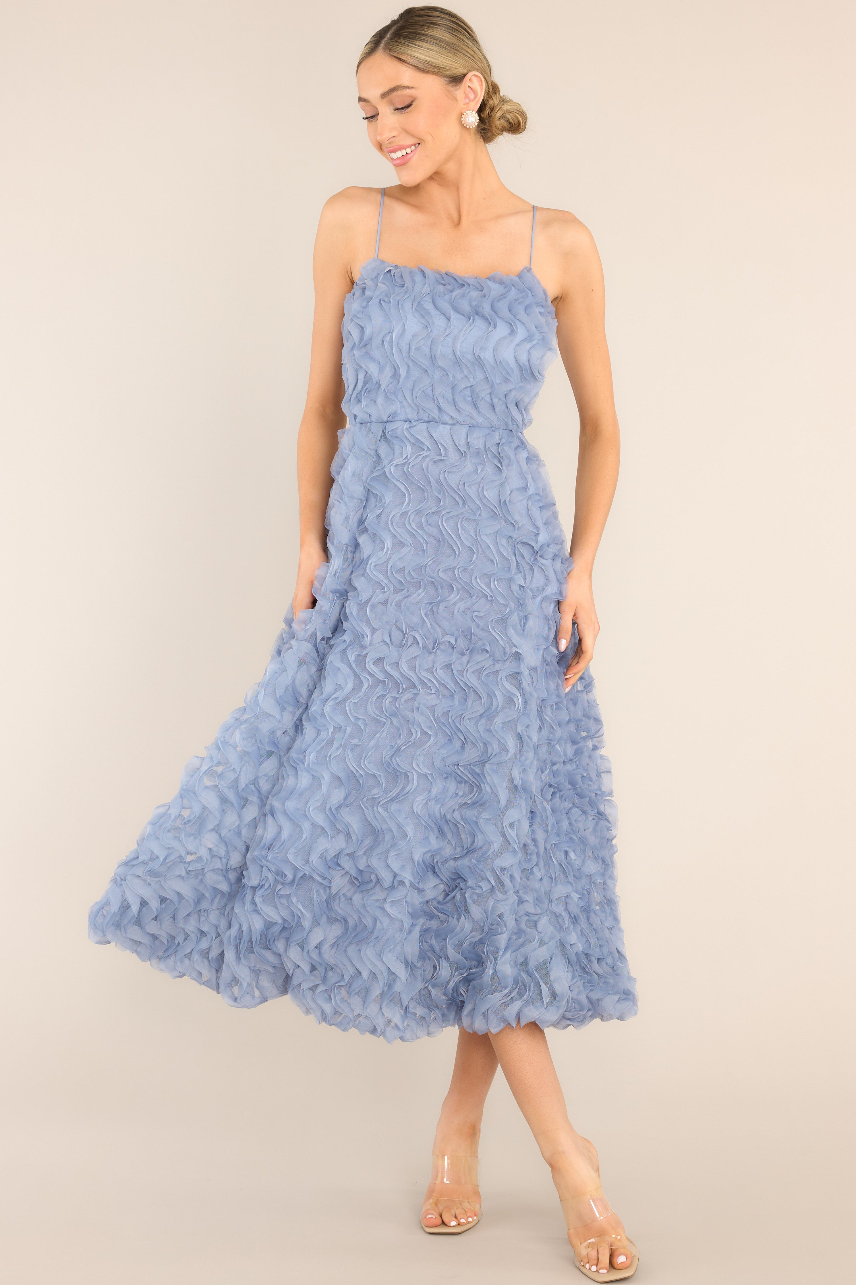 This blue dress features a rounded neckline, thin straps, a functional zipper down the back, and a wavy, ruffled overlay.