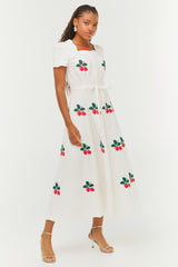 A model is shown wearing a white dress with embroidered red and green strawberries. The neckline and sleeve cuff are scalloped. There is a tie belt. She also wears silver heels with a strap.