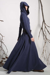 A woman wearing a long, blue hooded linen maxi dress with long sleeves, standing in an outdoor setting with trees in the background.
