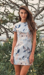 Outdoors, a woman wears the Mac Duggal Floral-Print Wedding-Guest Dress 11614 in light blue, with green foliage and tree branches behind her.