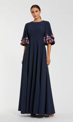 A woman models the Mac Duggal 11854 bell-sleeve long formal dress in navy blue, featuring pink and purple floral embellishments, set against a plain light background.