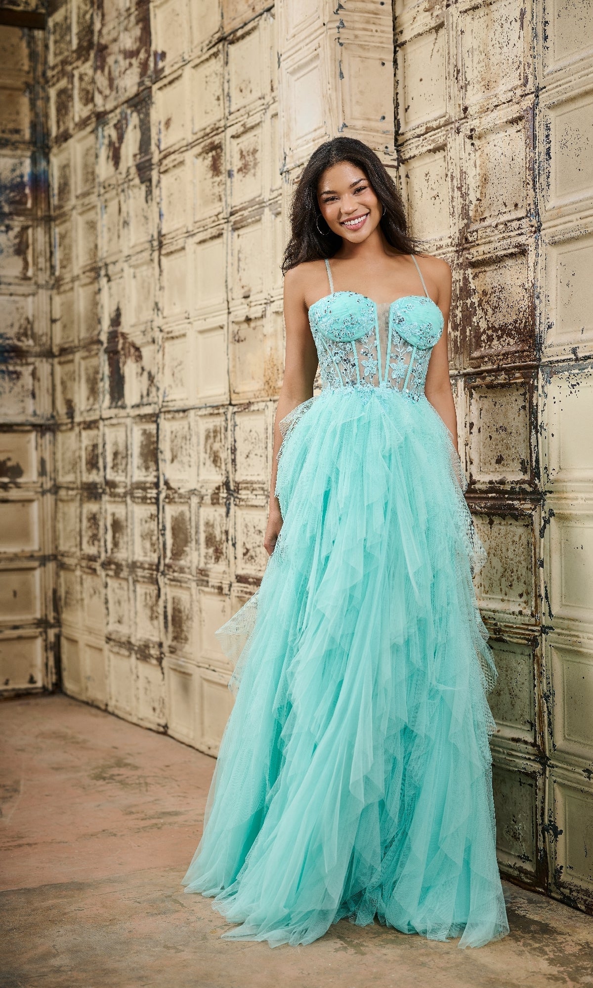 A woman stands before a textured beige wall wearing the Tiffany 16149 long prom dress—a light blue tulle gown with lace embroidery on the fitted bodice and a full, layered skirt.