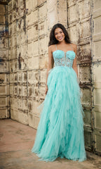 A woman stands before a textured beige wall wearing the Tiffany 16149 long prom dress—a light blue tulle gown with lace embroidery on the fitted bodice and a full, layered skirt.
