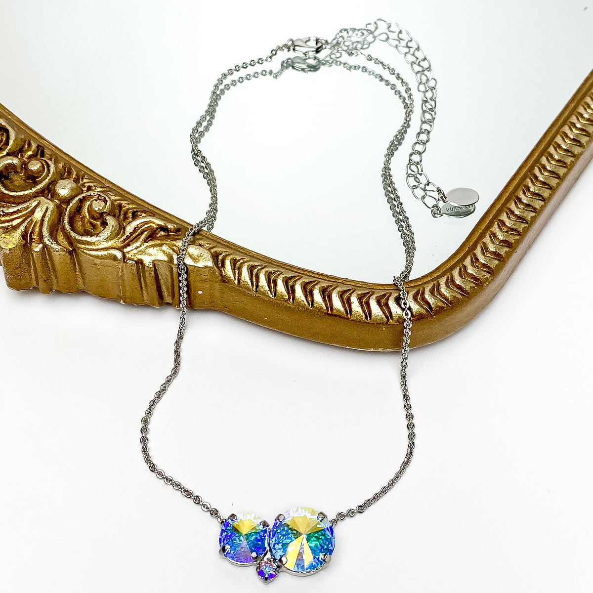 Pictured is a thin, silver chain necklace with a ab crystal pendant. The pendant is a cluster of three different sized, round crystals. This necklace is pictured partially laying on a gold mirror on a white background.