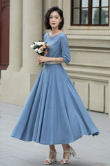 A woman wearing a blue fit and flare maxi swing linen dress with a boat neckline, half sleeves, and a back zipper closure.
