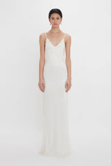 A woman in an elegant white bridal gown with crepe back satin, and lace detailing on the bodice, camisole-style straps, and a flowing skirt, stands against a plain white background. This stunning Exclusive Lace Detail Floor-Length Cami Dress In Ivory by Victoria Beckham is truly breathtaking.