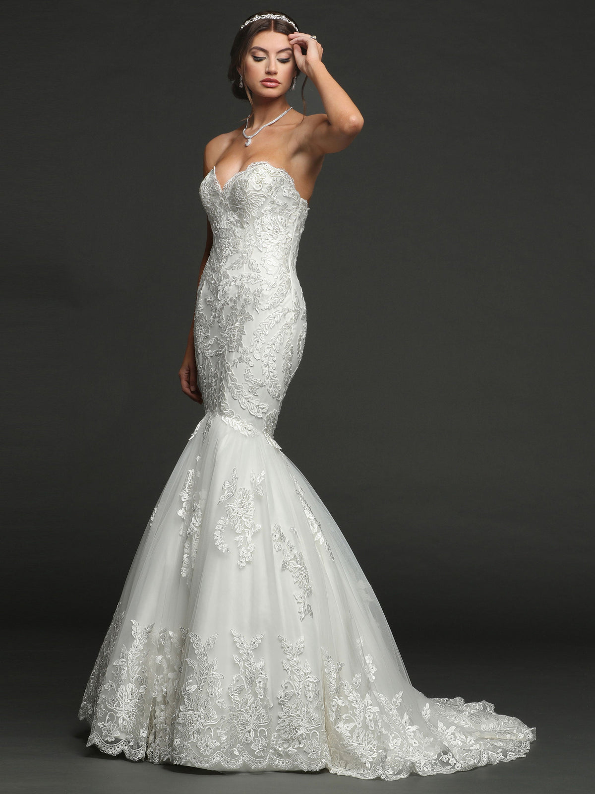 Wearing the Julia strapless mermaid wedding dress by Avery Austin, featuring lace details and a flared tulle skirt, a woman poses against a dark background with her hand softly touching her forehead.