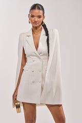 A woman with a long braided ponytail showcases the A LA MODE MINI BLAZER DRESS IN WHITE, featuring cape sleeves. She styles it with gold hoop earrings and a small gold clutch, posing confidently against a plain light background.