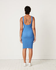 Wearing a fitted blue Hunza G Tank Dress and white sneakers, a short-haired individual stands with their back to us, revealing a large butterfly tattoo on their upper back in a simple, white-walled room.
