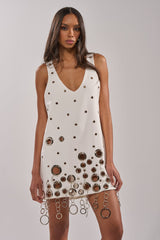A woman models the ONE OF A KIND MINI DRESS WITH METAL HARDWARE IN WHITE, featuring a deep V-neckline, metallic eyelets, and large metal rings with chain fringe along the hem, set against a plain background.