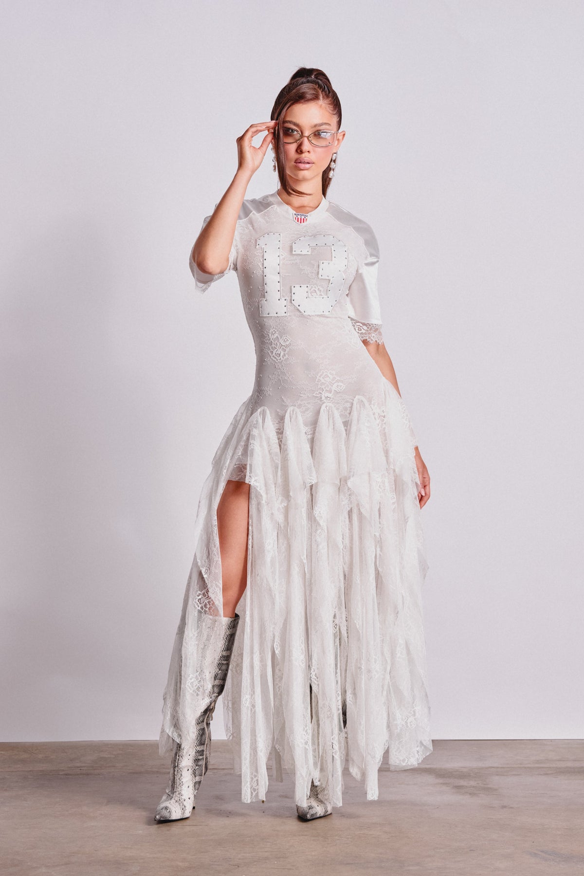Wearing the KICK OFF LACE JERSEY MAXI DRESS IN WHITE with a high slit, clear glasses, and knee-high patterned boots, this confident pose against a plain background is perfect festival outfit inspiration.