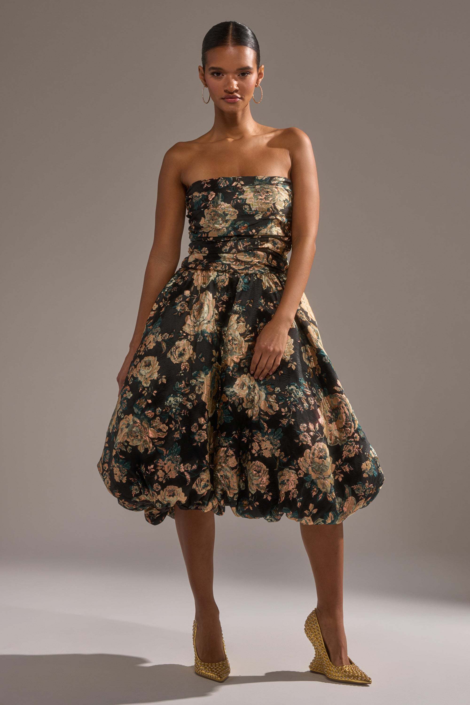 A woman poses confidently against a plain background in the DRESSED TO IMPRESS BROCADE MIDI DRESS, featuring a strapless bodice and voluminous skirt. She completes her look with gold hoop earrings, textured heels, and slicked-back hair.