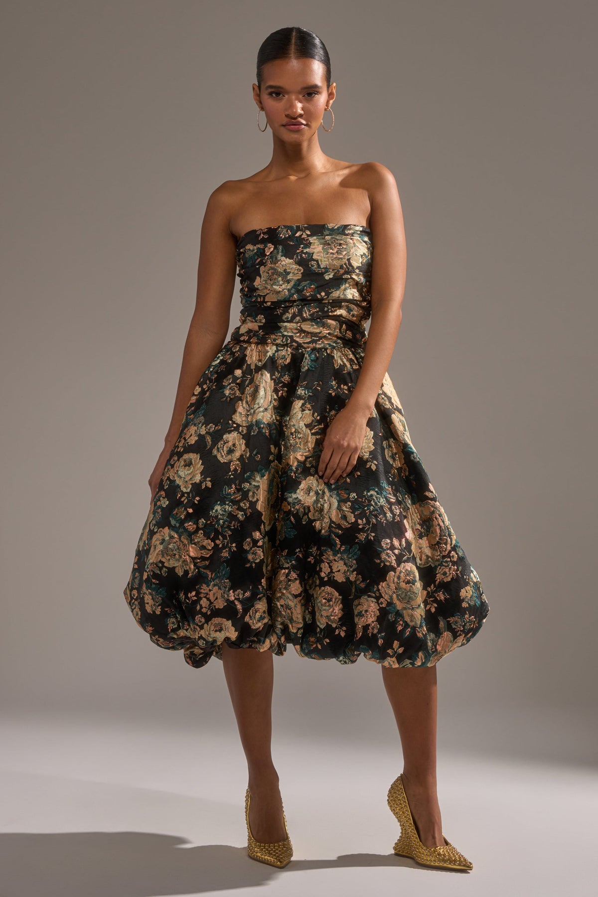 A woman poses confidently against a plain background in the DRESSED TO IMPRESS BROCADE MIDI DRESS, featuring a strapless bodice and voluminous skirt. She completes her look with gold hoop earrings, textured heels, and slicked-back hair.