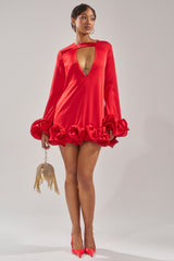 A woman wears the GIRLY GIRL SATIN MINI DRESS, a red mini with ruffled long sleeves, deep V neckline, and ruffled hem. She pairs it with red heels and a gold fringed handbag, standing against a plain light background.