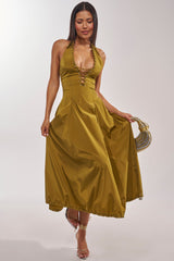 A woman in the SOMETHING LIGHT NYLON MAXI DRESS, featuring a mustard yellow halter-neck and A-line skirt, stands holding the sides of her dress. She accessorizes with strappy heels and a gold circular handbag.