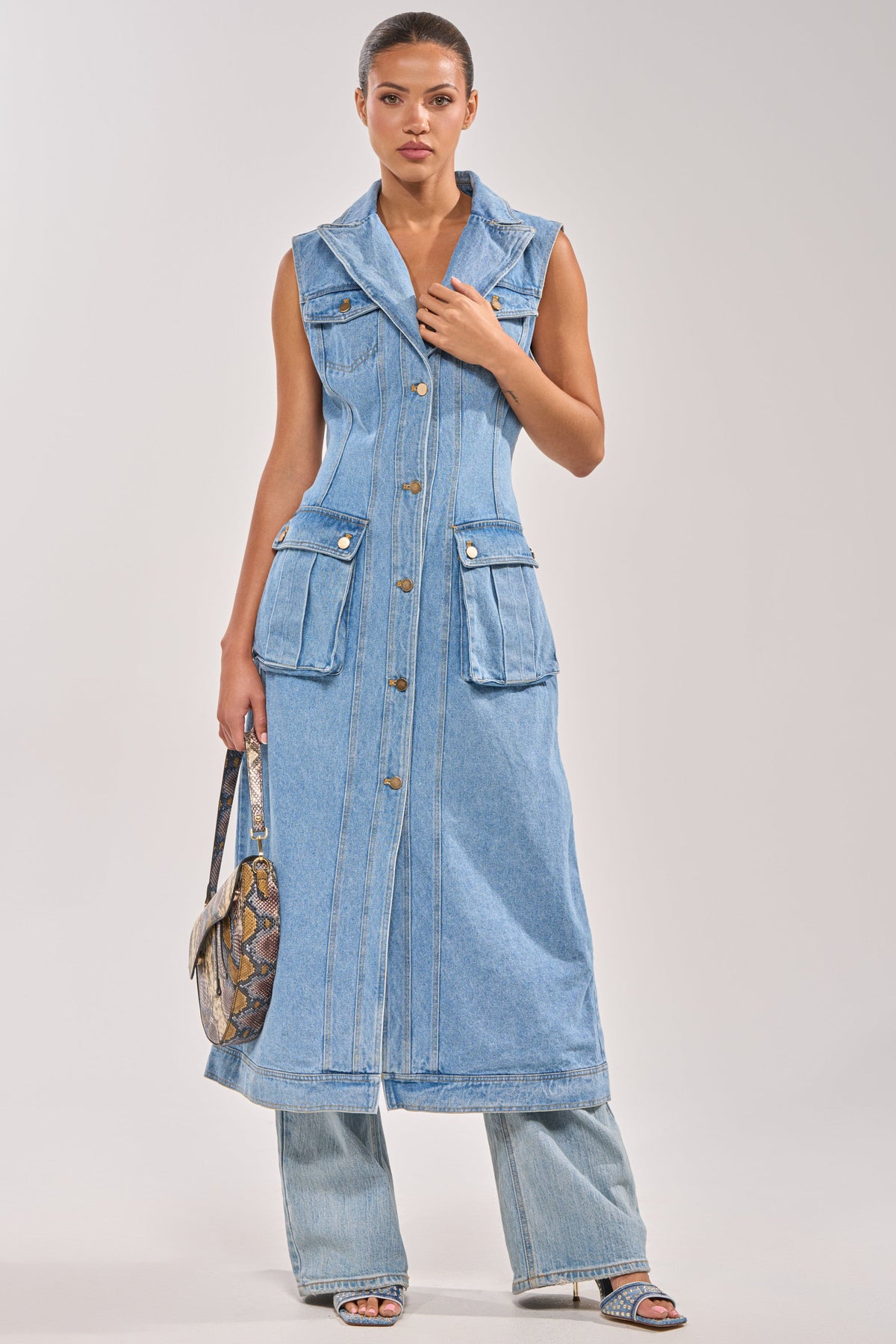 A woman faces forward wearing the GOTTA DIP LONG DENIM VEST, a sleeveless utility-inspired dress with front pockets, styled over wide-leg denim pants, open-toe heels, and a patterned handbag.