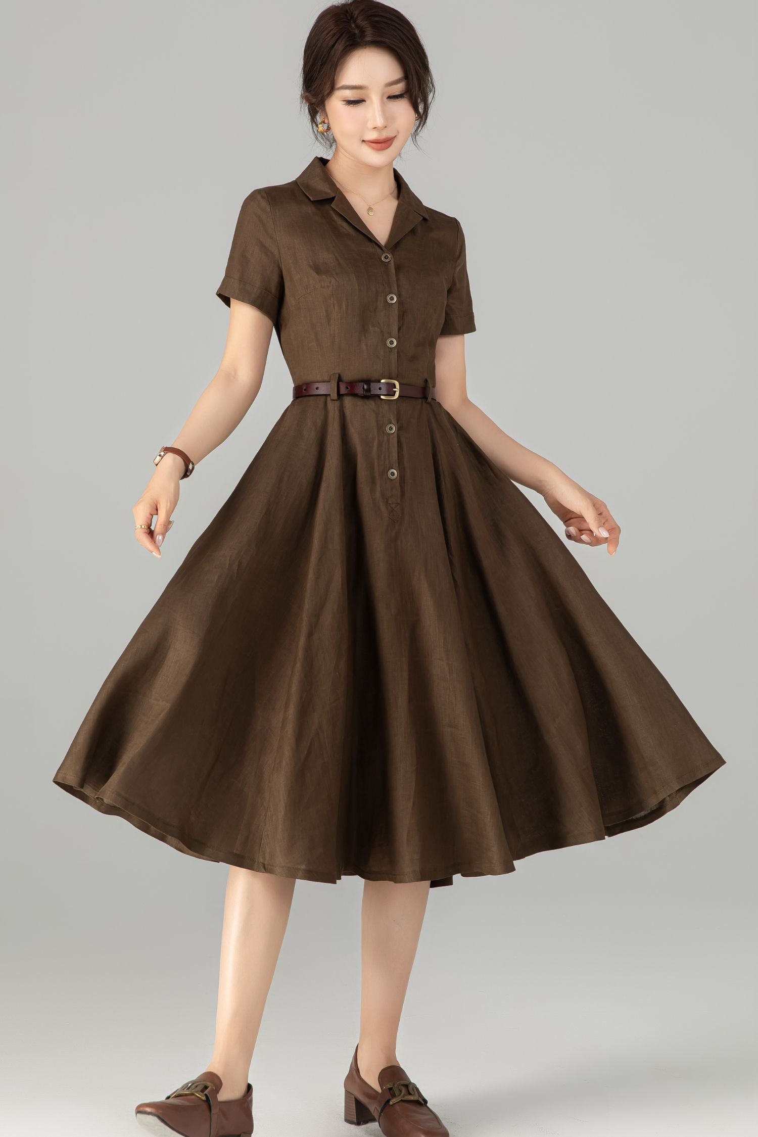 A woman wearing a brown button-up linen knee-length dress with short sleeves, paired with a belt and standing against a white background.