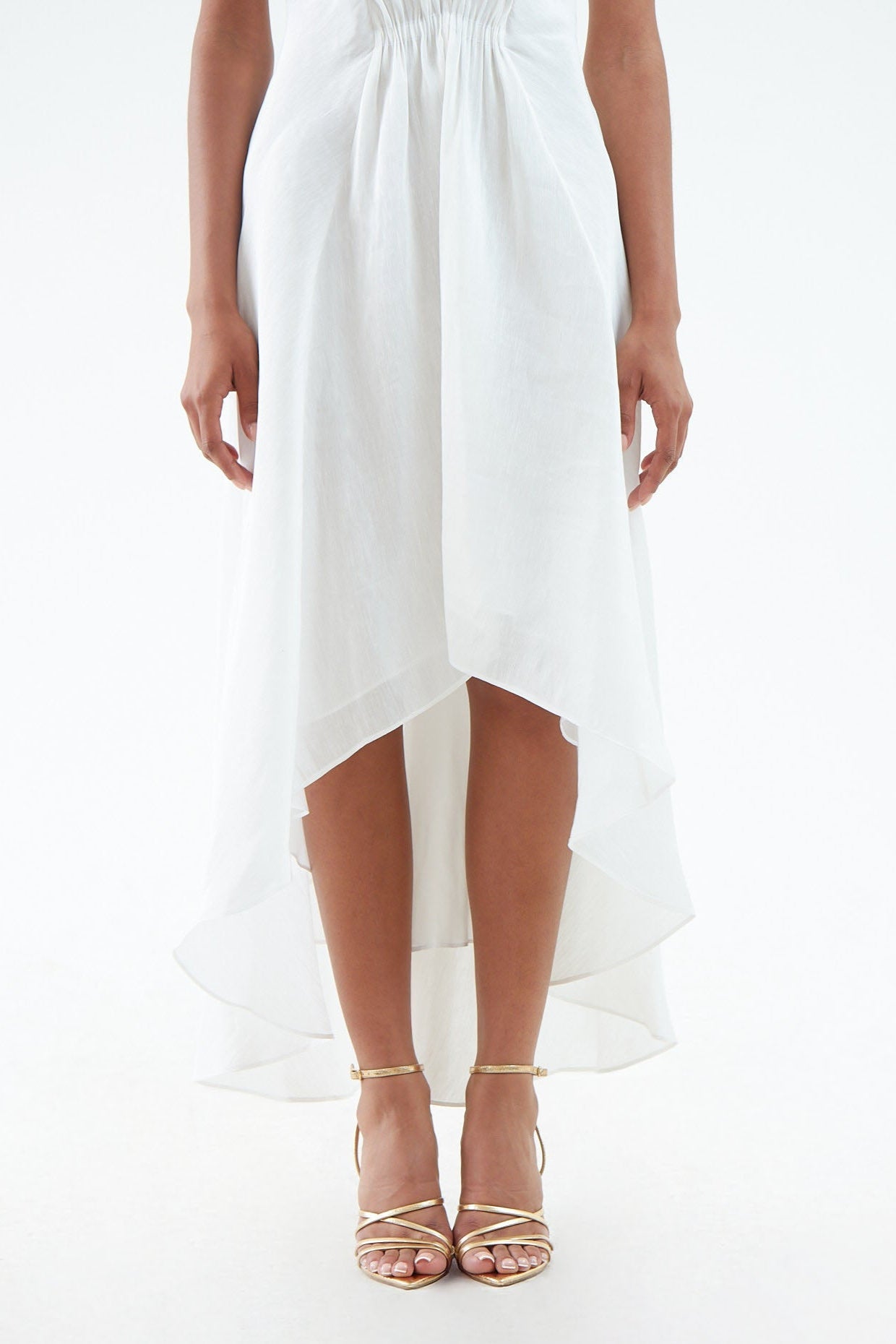 Zoomed in view of the lower portion of a white hemp silk dress, featuring a flowing short hem in the front and long hem in the back, and a gathered elastic waist. Paired with gold sandals. 
