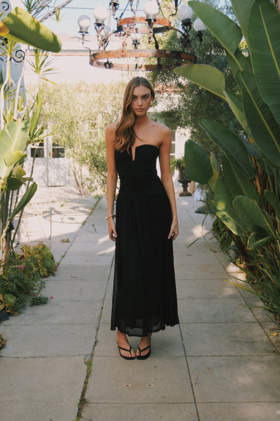 black-image | Woman in a black dress standing in a garden setting with plants and a chandelier.